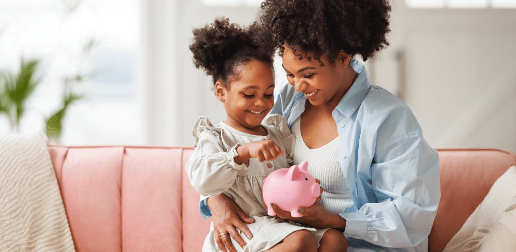 A mother and daughter sit on a pink couch, smiling as they put a coin into a pink piggy bank. The setting is bright and cozy, conveying warmth and bonding.