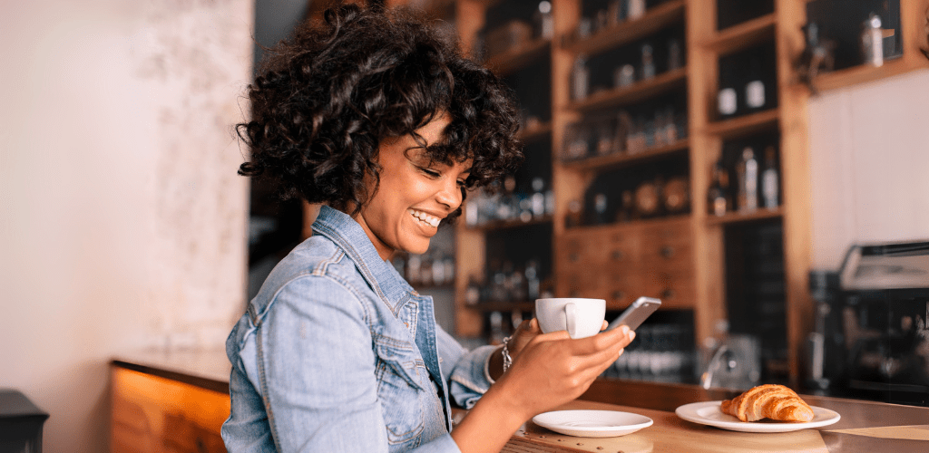 A woman in a denim jacket smiles while looking at her phone in a cozy cafe. She holds a cup of coffee, with a croissant on the counter. Relaxed atmosphere.
