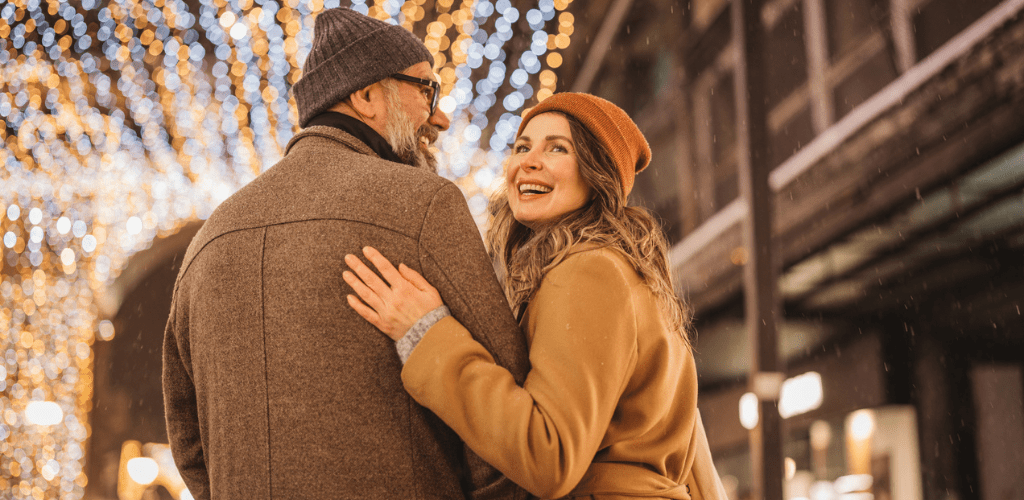 Couple wearing a coat and hat smiling under white holiday lights.